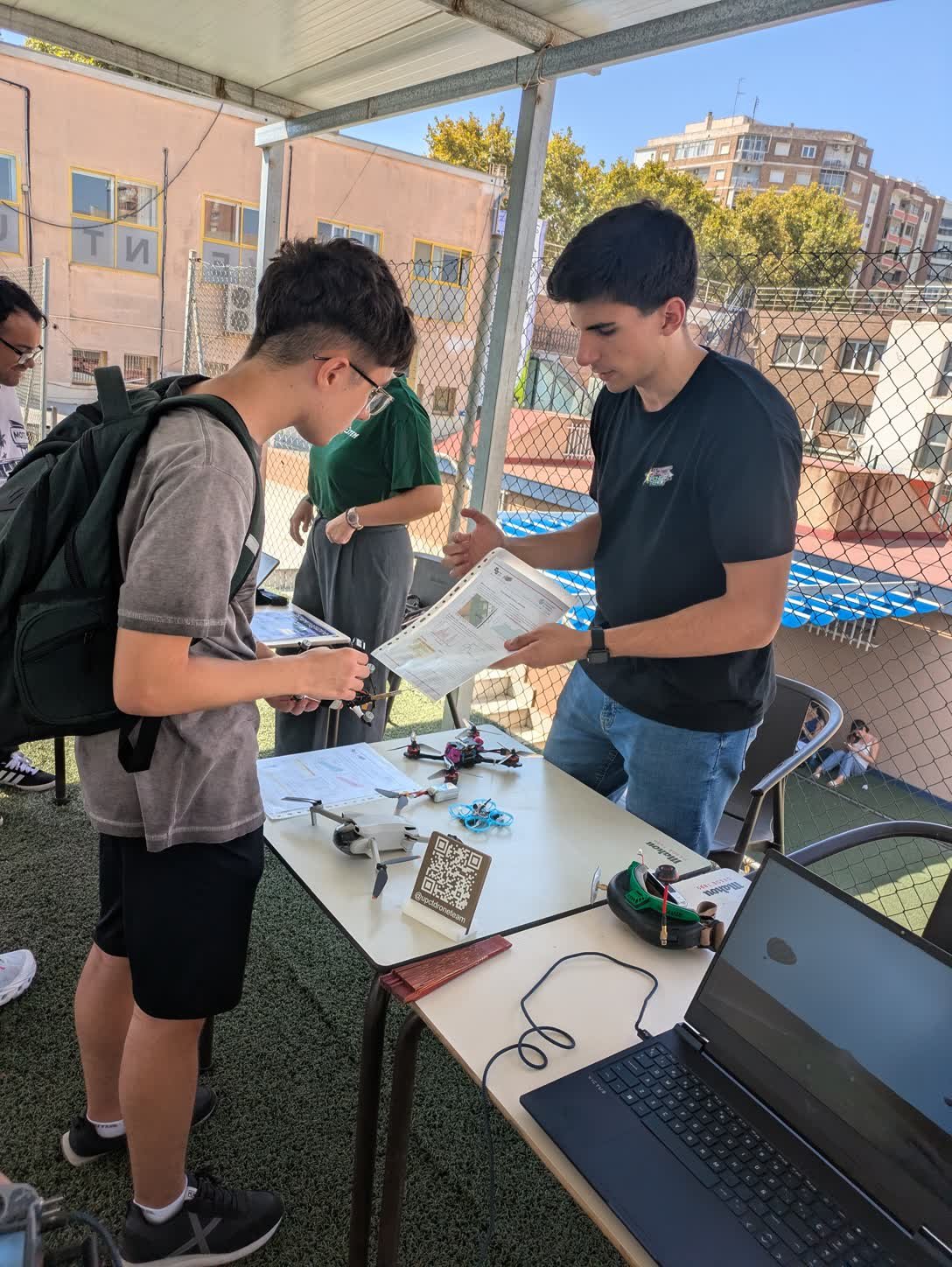 Un miembro del equipo explica el material a un visitante que sostiene la emisora, con el dron FPV, el DJI Mini, el tinywhoop y las gafas sobre la mesa