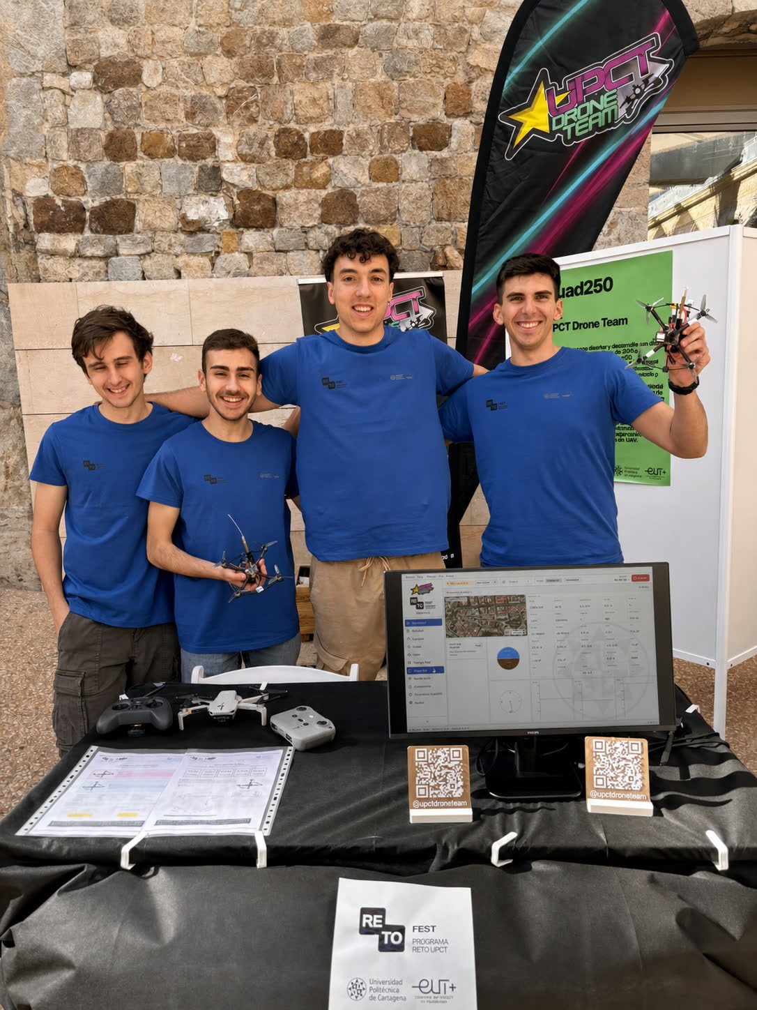 Los cuatro miembros del equipo en el stand del RetoFest, uno sosteniendo el Quad250, con la telemetría en un monitor y el cartel del reto detrás