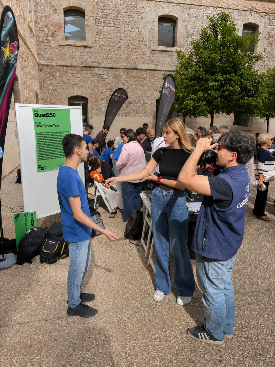 Un miembro del equipo entrevistado por una televisión junto al cartel del Reto Quad250 en el RetoFest